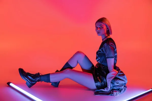 Stylische Frau in Minirock und Lederstiefeln, die neben lila Neonlampen auf korallenrotem Hintergrund sitzt — Stockfoto