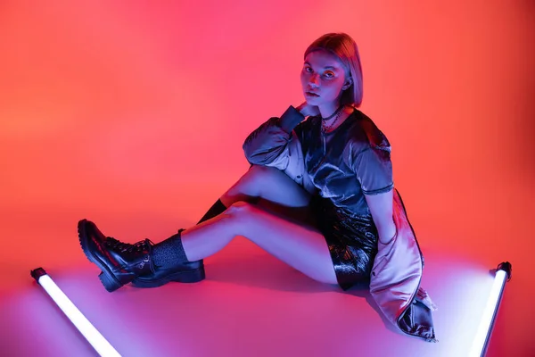 Full length of woman in leather boots and mini skirt looking at camera near neon lamps on purple and coral background — Stock Photo