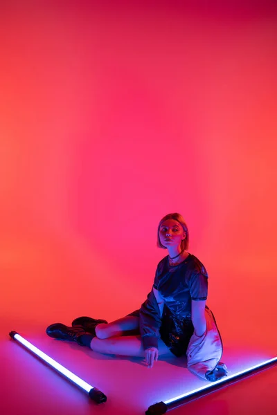 Full length of stylish woman sitting near purple neon lamps on coral red background — Stock Photo