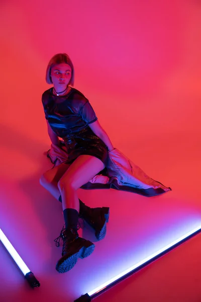 Vista de alto ângulo da mulher em botas de couro preto sentado perto de lâmpadas de néon brilhantes no fundo roxo e coral — Fotografia de Stock