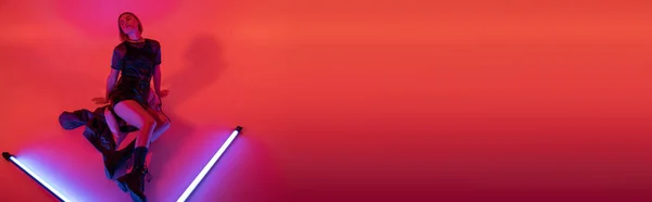 Високий кут зору молодої жінки в чоботях і міні-спідниці позує біля сяючих неонових ламп на фіолетовому і червоному тлі, банер — стокове фото