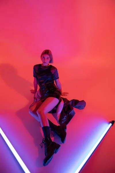 High angle view of woman in mini skirt and leather boots sitting near purple neon lamps on coral red background — Stock Photo