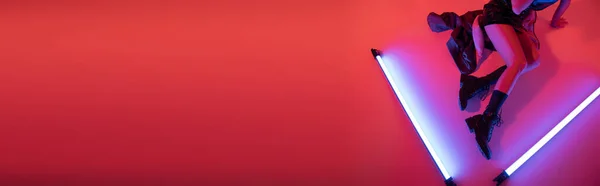 Vista superior da mulher cortada em botas sentado perto de lâmpadas de néon roxo no fundo vermelho carmim, banner — Fotografia de Stock