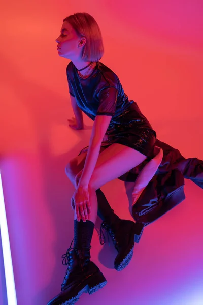 Vista de alto ângulo da mulher em mini-saia preta e botas de couro sentado perto de lâmpada de néon no fundo roxo e coral — Fotografia de Stock