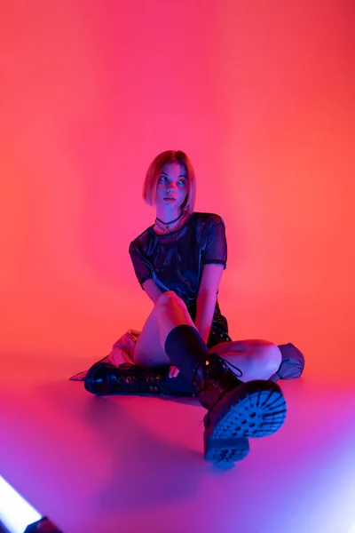 Молода жінка в стильних шкіряних чоботях, дивлячись далеко, сидячи в неоновому світлі на фіолетовому і кораловому фоні — стокове фото