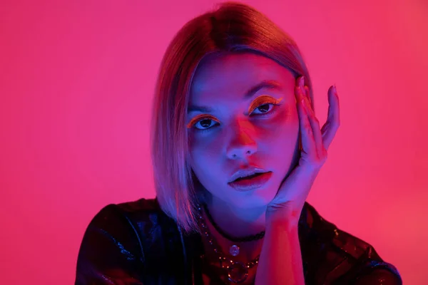 Retrato de mulher bonita com maquiagem de néon segurando mão perto do rosto em luz azul no fundo rosa profundo — Fotografia de Stock