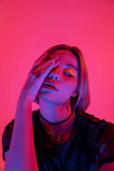 Mulher com maquiagem de néon e colares obscurecendo o rosto com a mão em luz roxa no fundo rosa profundo — Fotografia de Stock