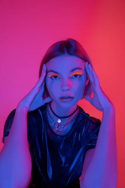 Trendfrau in hellem Neon-Make-up und Halsketten posiert mit händennahem Gesicht auf dunkelrosa und korallenfarbenem Hintergrund — Stockfoto