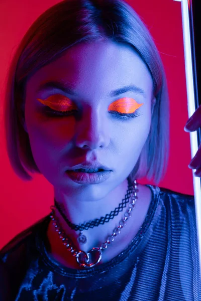 Portrait of woman with neon makeup in blue light of glowing lamp on carmine red background — Stock Photo