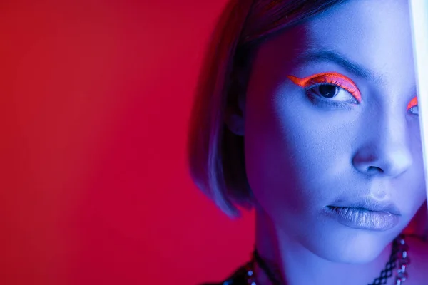 Vue recadrée de la femme avec du maquillage au néon regardant la caméra en lumière bleue sur fond rouge carmin — Photo de stock