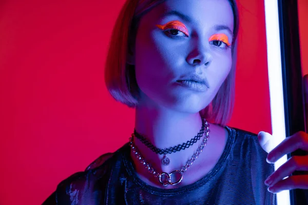 Trendy woman in bright makeup and necklaces looking at camera in blue light of neon lamp on carmine red background — Stock Photo
