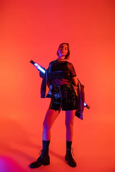 Volle Länge der trendigen Frau in Minirock und schwarzen Stiefeln posiert mit Neon-Lampe auf korallenrotem Hintergrund — Stockfoto