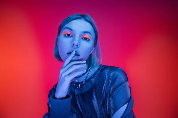 Sensual young woman with neon makeup touching lips in blue light on coral and pink background — Stock Photo