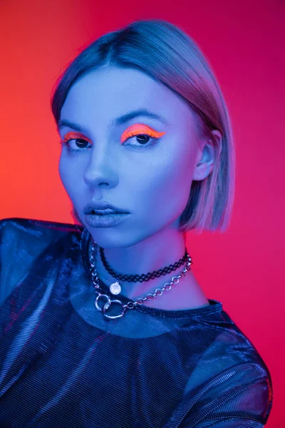 Mulher na moda em colares e maquiagem de néon olhando para a câmera em luz azul no fundo coral e rosa — Fotografia de Stock