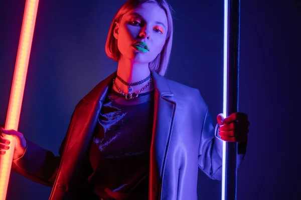 Trendy woman in leather jacket holding neon lamps while looking at camera on dark blue background — Stock Photo