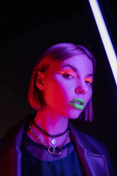 Trendige Frau mit hellem Neon-Make-up, die in die Kamera in der Nähe lila Leuchtstofflampe auf schwarzem Hintergrund schaut — Stockfoto