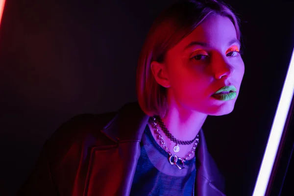 Junge Frau mit Neon-Make-up blickt in die Kamera im lila Licht der Leuchtstofflampe auf schwarzem Hintergrund — Stockfoto