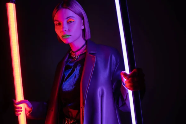 Woman in leather jacket and neon makeup holding glowing fluorescent lamps on dark purple background — Stock Photo