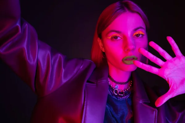 Donna in giacca di pelle e vivace trucco al neon guardando la fotocamera su sfondo viola scuro — Foto stock