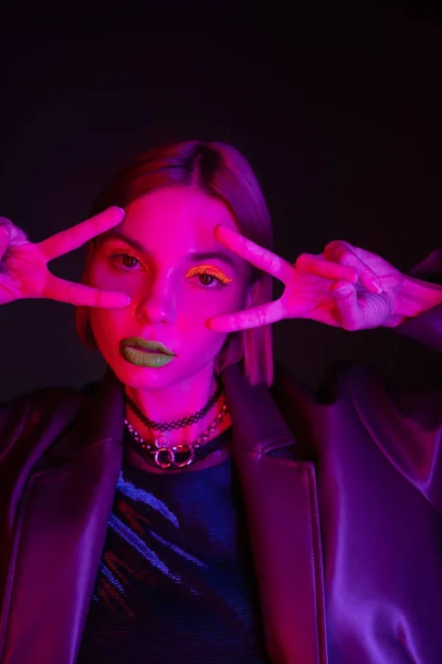 Mulher na moda em maquiagem de néon e colares mostrando sinais de vitória e olhando para a câmera em luz roxa no fundo preto — Fotografia de Stock
