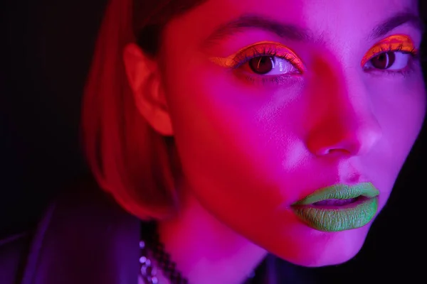 Close up portrait of woman with glowing neon makeup in purple light on black background — Stock Photo