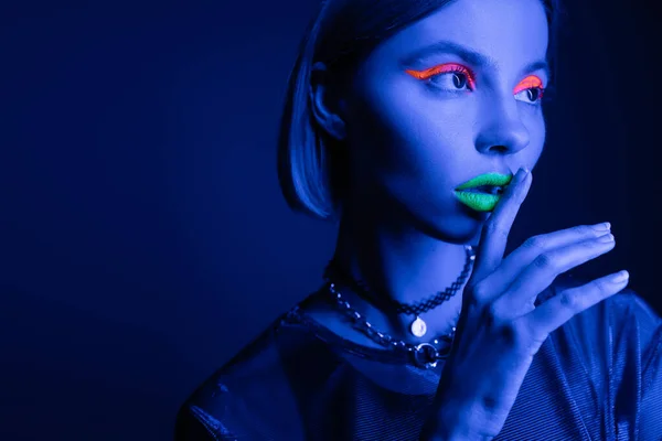Retrato de mulher em maquiagem de néon tocando lábios verdes enquanto mostra sinal de silêncio no fundo azul escuro — Fotografia de Stock