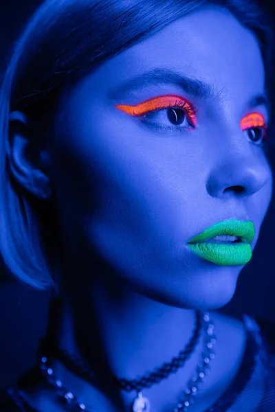 Close up portrait of woman with vibrant makeup looking away in neon light on dark blue background — Stock Photo