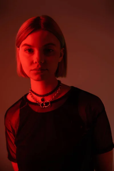 Молодая и модная женщина в ожерельях смотрит в камеру в красном свете на бордовом фоне — стоковое фото