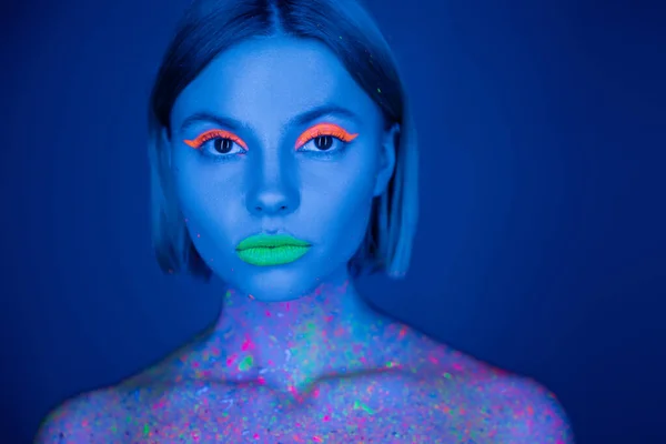 Портрет женщины в ярком неоновом макияже и флуоресцентной краске тела, смотрящей на камеру, изолированную на темно-синем — стоковое фото