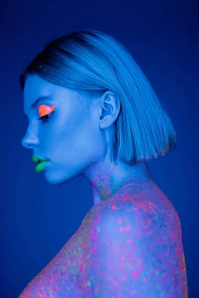 Profil de jeune femme avec maquillage au néon et éclaboussures de peinture brillantes sur fond bleu — Photo de stock