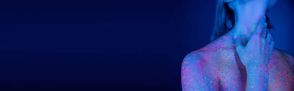 Vista ritagliata di donna nuda con brillanti spruzzi al neon sul corpo toccando il collo su sfondo blu scuro, banner — Foto stock