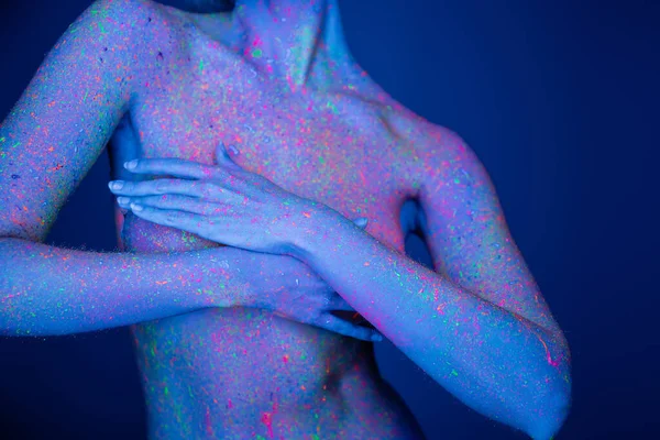 Vista cortada de mulher nua com salpicos de néon brilhante no corpo cobrindo peito com as mãos isoladas no azul escuro — Fotografia de Stock