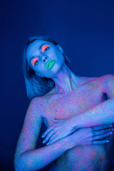 Mujer desnuda con maquillaje vibrante y pintura de neón en el cuerpo aislado en azul oscuro — Stock Photo