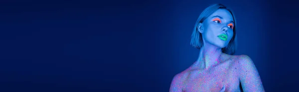 Nude woman in glowing neon makeup and colorful paint splashed looking away on dark blue background, banner — Stock Photo