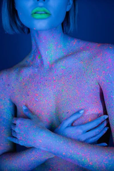 Vue recadrée d'une femme nue avec des lèvres au néon vertes et des éclaboussures de peinture brillantes sur le corps couvrant la poitrine isolée sur bleu foncé — Photo de stock
