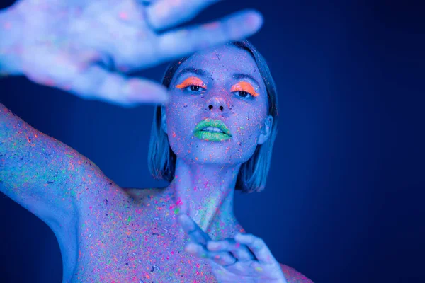 Frau in leuchtendem Make-up und Neon-Körperfarbe posiert mit verschwommener ausgestreckter Hand isoliert auf dunkelblau — Stockfoto