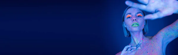 Жінка в яскравій неоновій фарбі для тіла і макіяж позує з розмитою простягнутою рукою на темно-синьому фоні, банер — стокове фото
