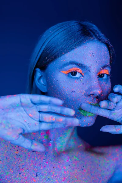 Junge Frau mit Neon-Make-up und bunten Farbspritzern posiert mit Händen in Gesichtsnähe isoliert auf dunkelblau — Stockfoto