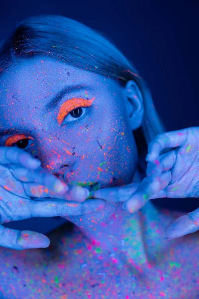 Young woman in fluorescent paint and neon makeup holding hands near face isolated on dark blue — Stock Photo