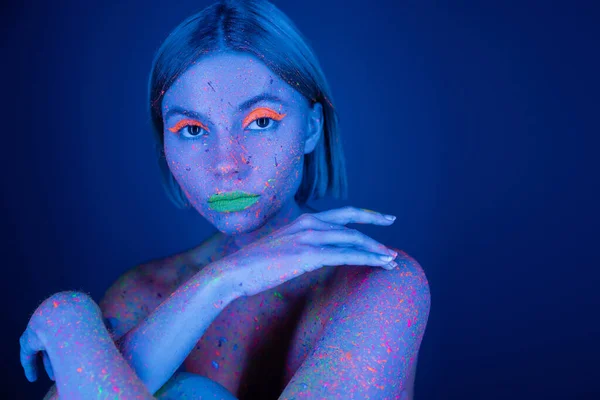 Оголена жінка в яскравій фарбі для тіла і неоновий макіяж дивиться на камеру ізольовано на темно-синьому — стокове фото