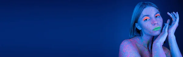 Junge Frau in heller Neon-Körperfarbe und leuchtendem Make-up, die isoliert auf dunkelblauem Banner in die Kamera blickt — Stockfoto
