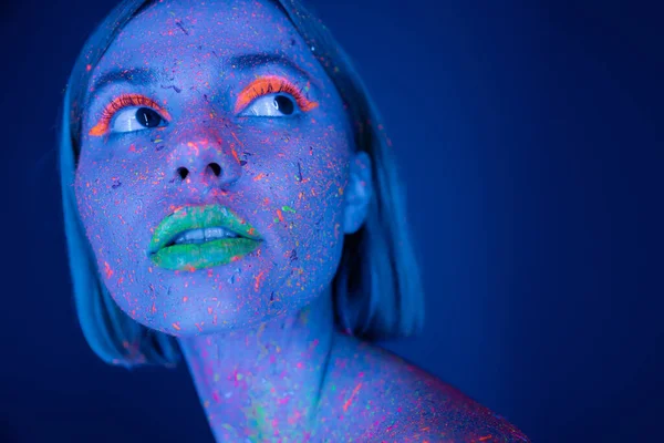 Portrait of young woman with neon makeup and vibrant paint on face looking away isolated on dark blue — Stock Photo