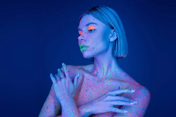 Nackte Frau in Neon-Make-up und glühender Farbe spritzt auf Körper posiert mit verschränkten Armen isoliert auf blau — Stockfoto