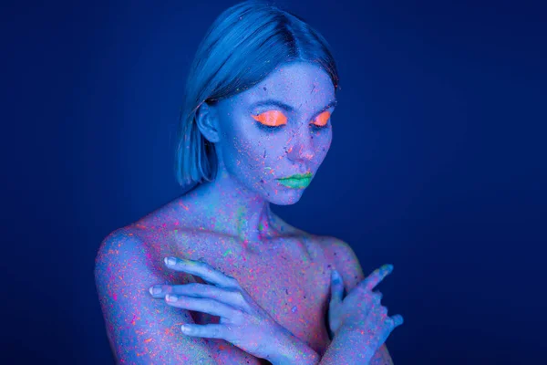 Mulher nua em maquiagem de néon brilhante e pintura corporal brilhante cobrindo o peito com as mãos isoladas no azul escuro — Fotografia de Stock