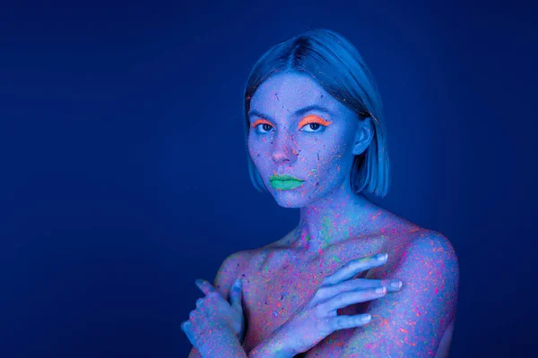 Mulher nua com maquiagem vibrante e corpo em tinta de néon olhando para a câmera isolada em azul escuro — Fotografia de Stock