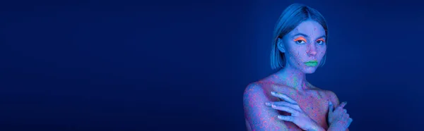 Mulher em maquiagem neon brilhante e respingos de tinta coloridos posando com braços cruzados no fundo azul escuro, banner — Fotografia de Stock