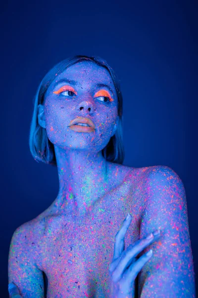 Mulher nua com corpo colorido e maquiagem neon brilhante olhando para longe isolado no azul escuro — Fotografia de Stock