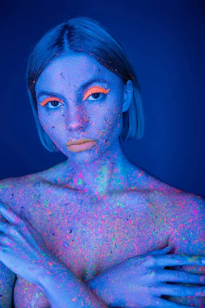 Nude woman with neon makeup and bright paint on body covering bust with hands isolated on dark blue — Stock Photo