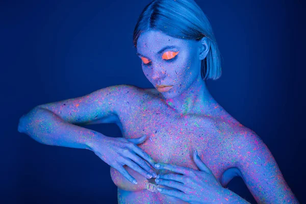 Jovem mulher em respingos de tinta brilhante e maquiagem de néon cobrindo o peito com as mãos isoladas no azul escuro — Fotografia de Stock