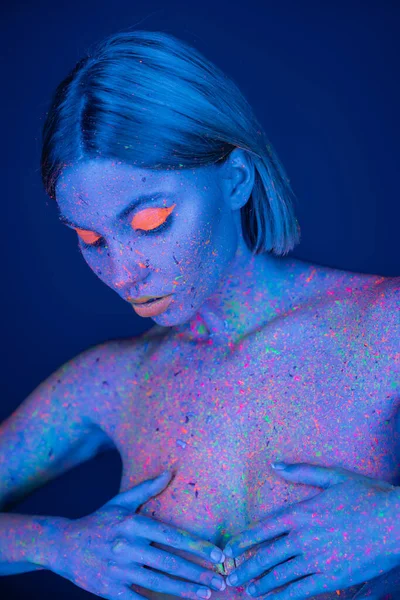 Молода і гола жінка з сяючим макіяжем і кольоровим тілом, що покриває груди руками ізольовані на темно-синьому — стокове фото